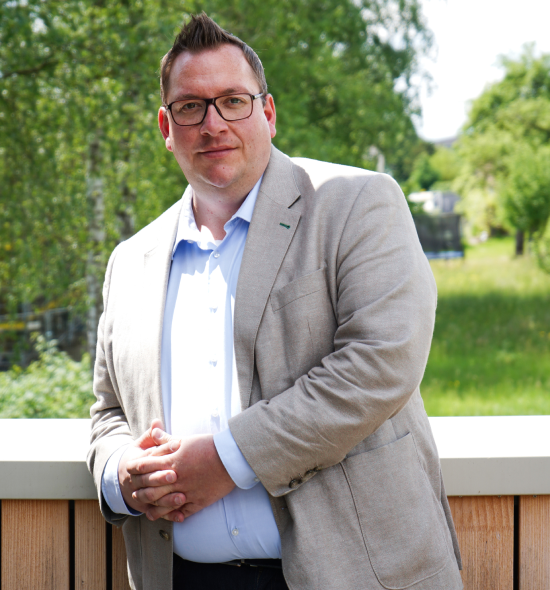 Mann in einem Anzug lehnt an einem Geländer mit grüner Umgebung im Hintergrund, er lächelt und schaut in die Kamera.