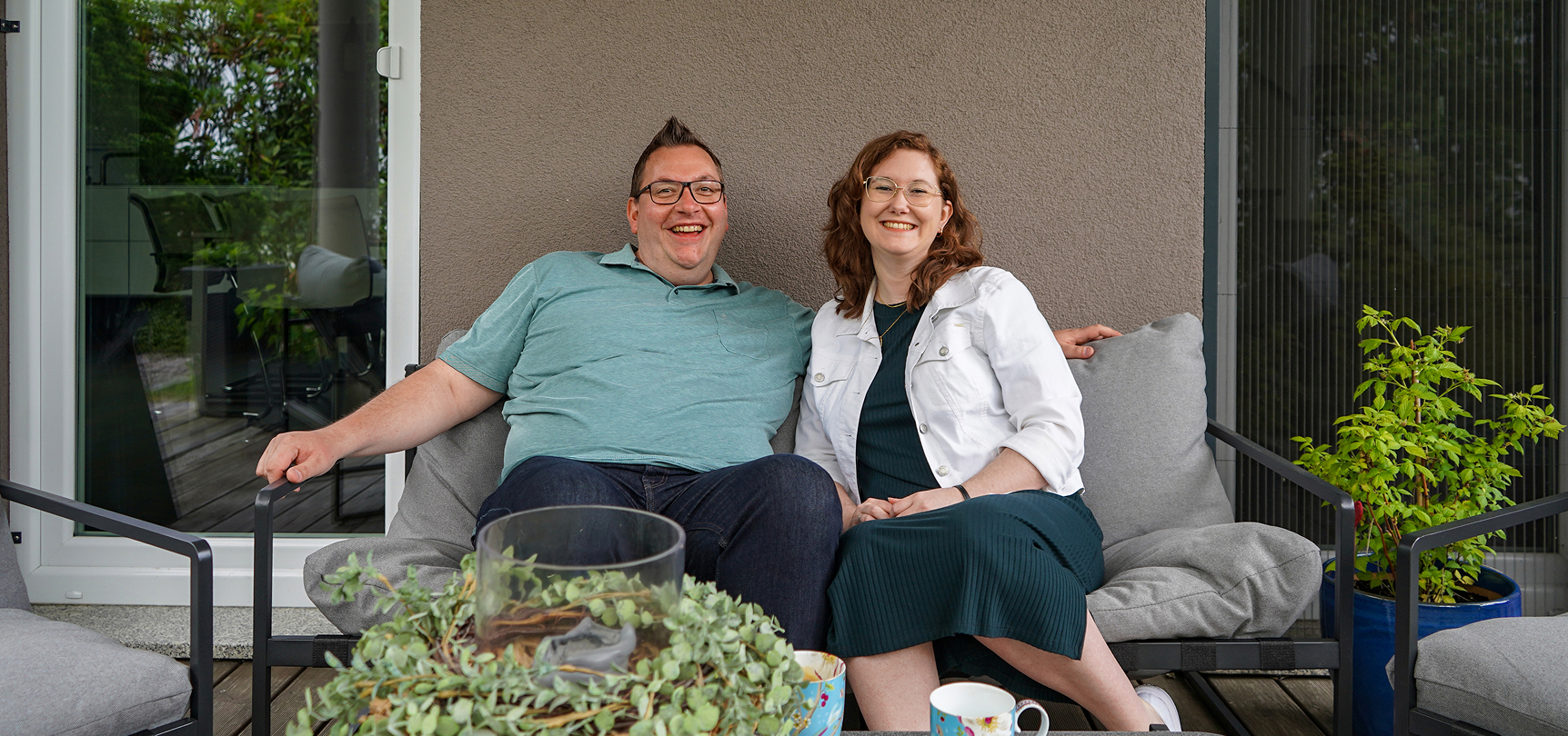 Ein Paar sitzt entspannt auf einer Terrasse, umgeben von Pflanzen und Tassen. Beide lächeln in die Kamera.