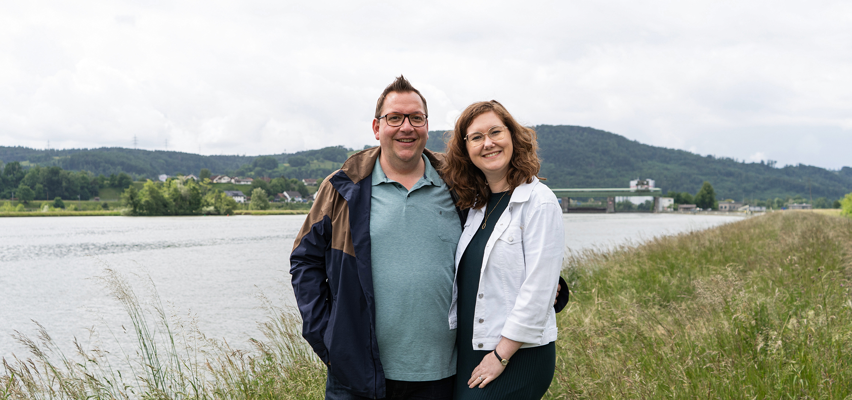 Ein Paar steht am Ufer eines Gewässers, umgeben von Wiesen und sanften Hügeln, lächeln in die Kamera.