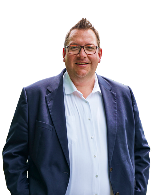 Mann mit Brille, fröhlich lächelnd, trägt ein weißes Hemd und einen dunklen Blazer, vor einem einfarbigen Hintergrund.